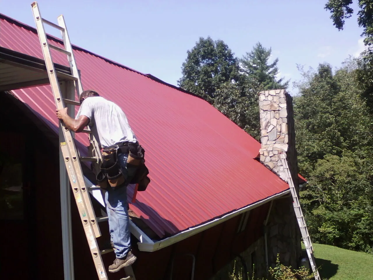 Roofer on a ladder beside a red metal roof and stone chimney — Soffit & Fascia Repair by Roofing Brothers in North Powder